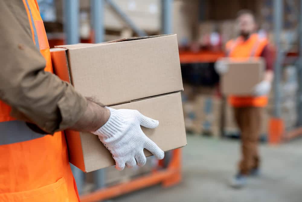 Warehouse worker in orange safety vest carrying cardboard boxes in distribution center