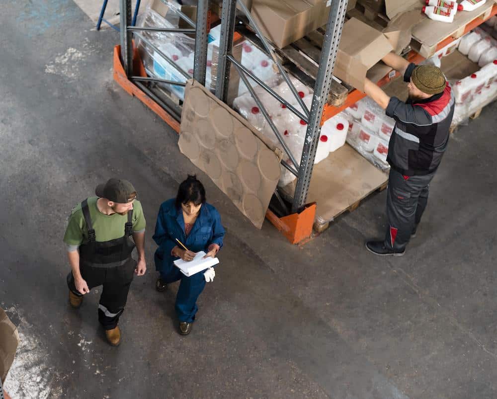 Three warehouse workers conducting inventory inspection with pallet jack and storage shelves