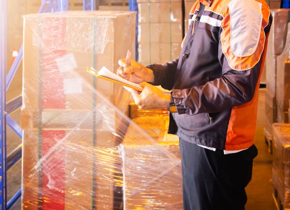 Warehouse worker with clipboard managing inventory among stacked boxes and packages