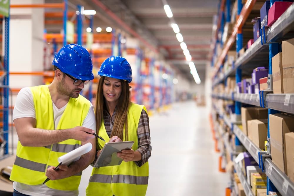 Two warehouse workers in safety gear reviewing inventory on tablet and clipboard