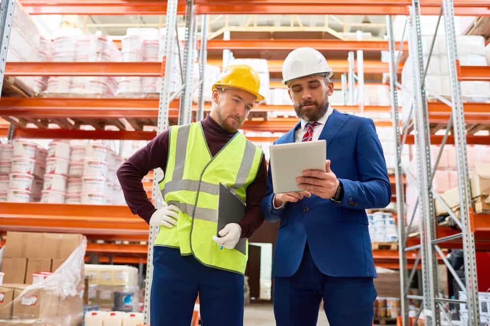 Two businessmen reviewing tablet data in industrial warehouse with orange shelving