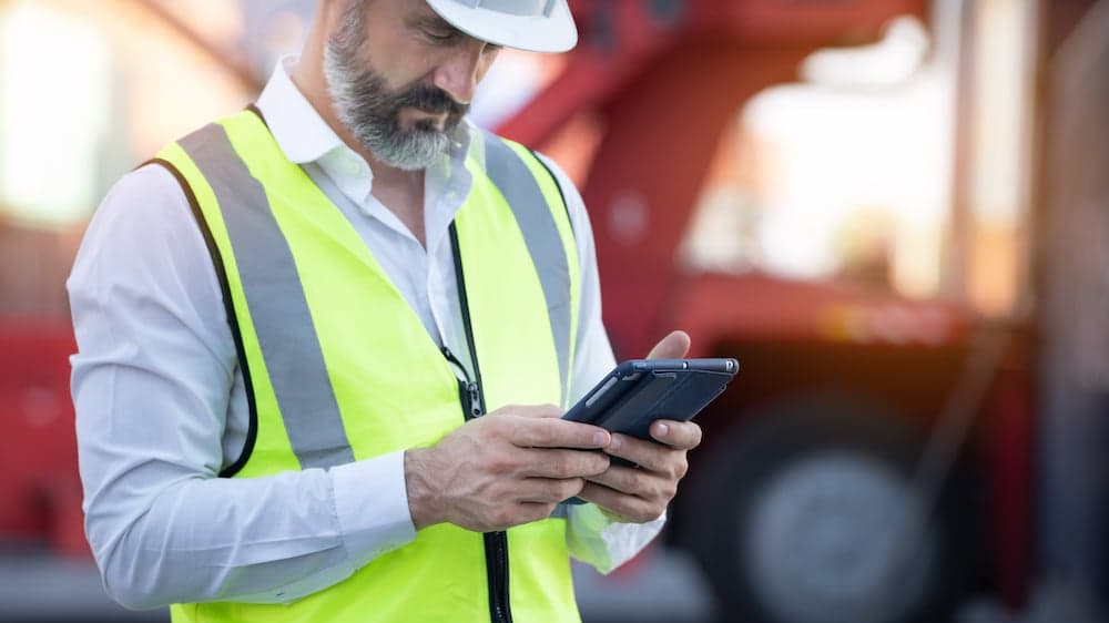 Manager in safety vest and hard hat using tablet at shipping container terminal