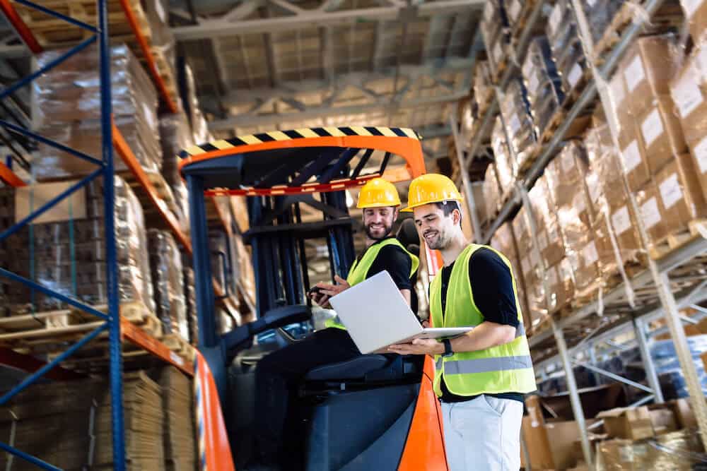 Two warehouse workers in safety gear reviewing documents near forklift in distribution center