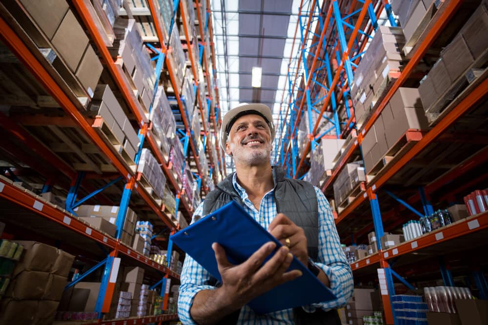 Warehouse worker with hard hat checking inventory on clipboard in storage facility