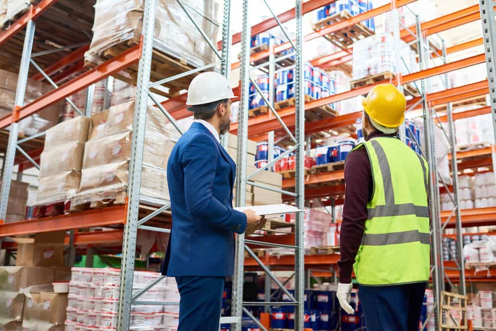 Two warehouse workers in hard hats inspecting inventory on industrial shelving