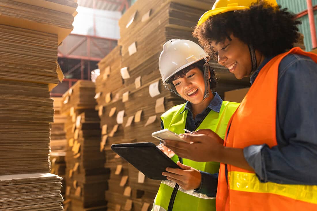 Two female warehouse workers in safety gear reviewing inventory on tablet and smartphone