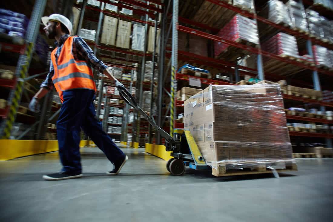 Warehouse worker in safety vest pulling pallet jack with boxes in distribution center