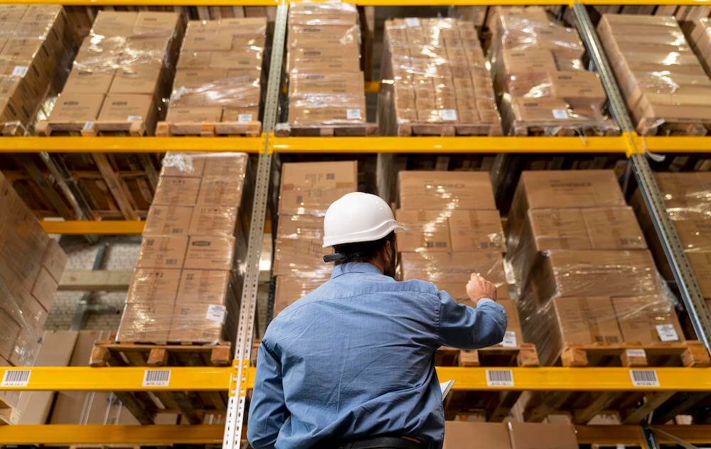 Warehouse worker in white hard hat inspecting inventory on storage shelves