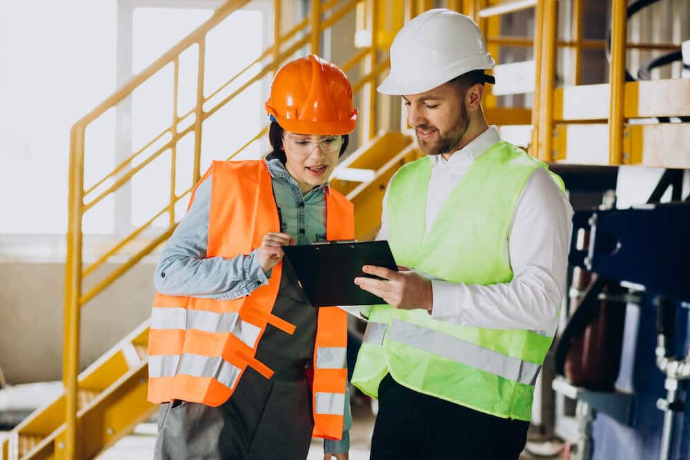 Two factory workers in safety gear reviewing clipboard documents together