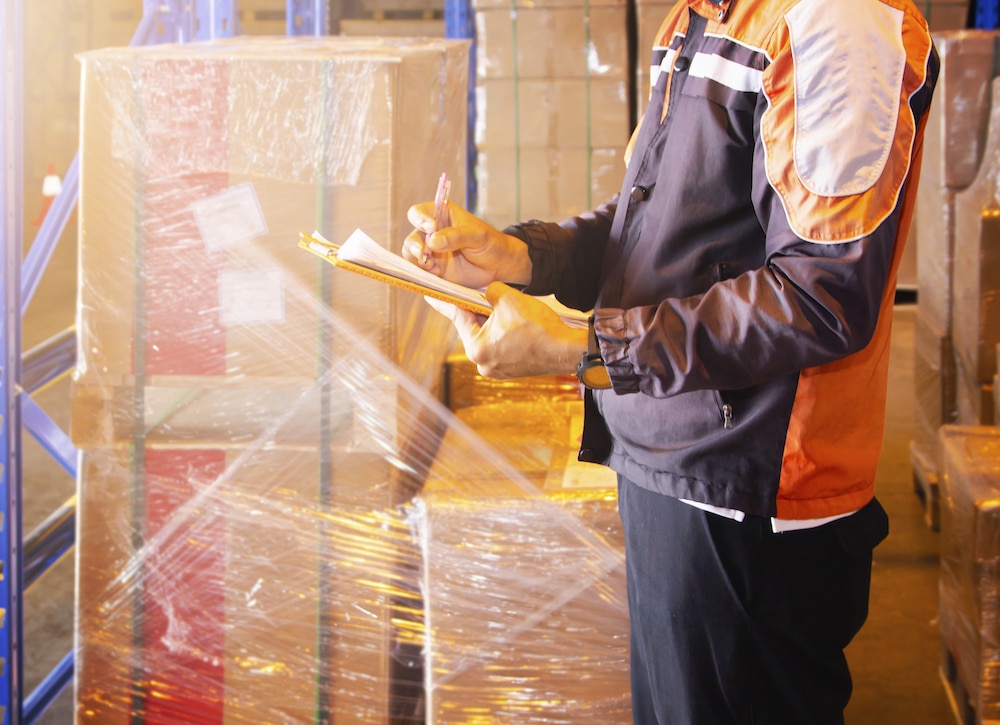 Warehouse worker with clipboard managing inventory among stacked boxes and packages