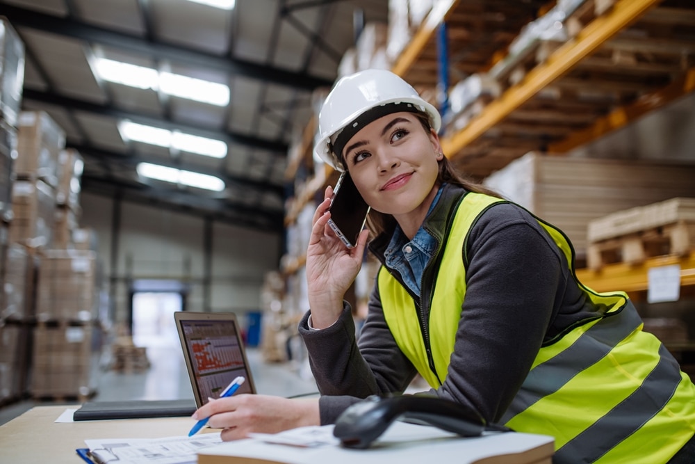 warehouse worker on a cell phone at a desk with a laptop and scanner