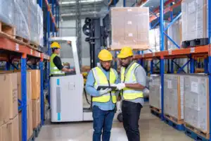2 warehouse workers looking at a tablet and behind them something moving a pallet on a forklift