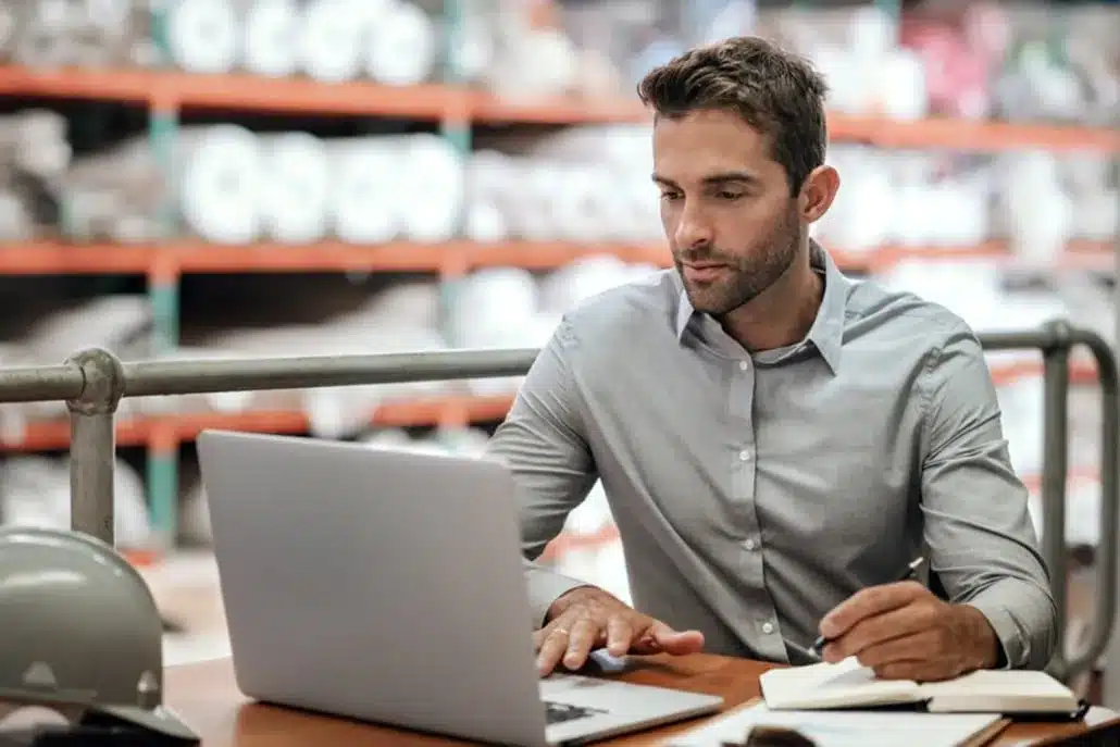 a man at a desk looking at a laptop with warehouse shelves in the background