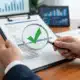 Hands examining clipboard with magnifying glass showing checkmark, representing supply chain efficiency analysis